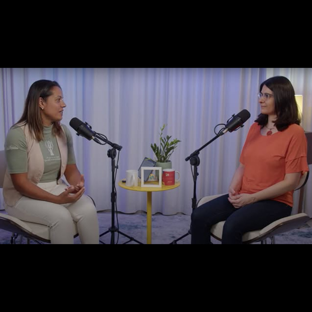 Vânia Ferreira e Cláudia Nascimento conversam com microfones uma de frente para a outra. Elas estão sentadas em uma sala com cortina ao fundo e uma mesinha amarela ao centro.
