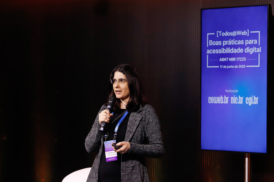 Destaque de Cláudia Nascimento falando ao microfone. Ela veste blazer cinza por cima de malha preta e usa um crachá do evento.