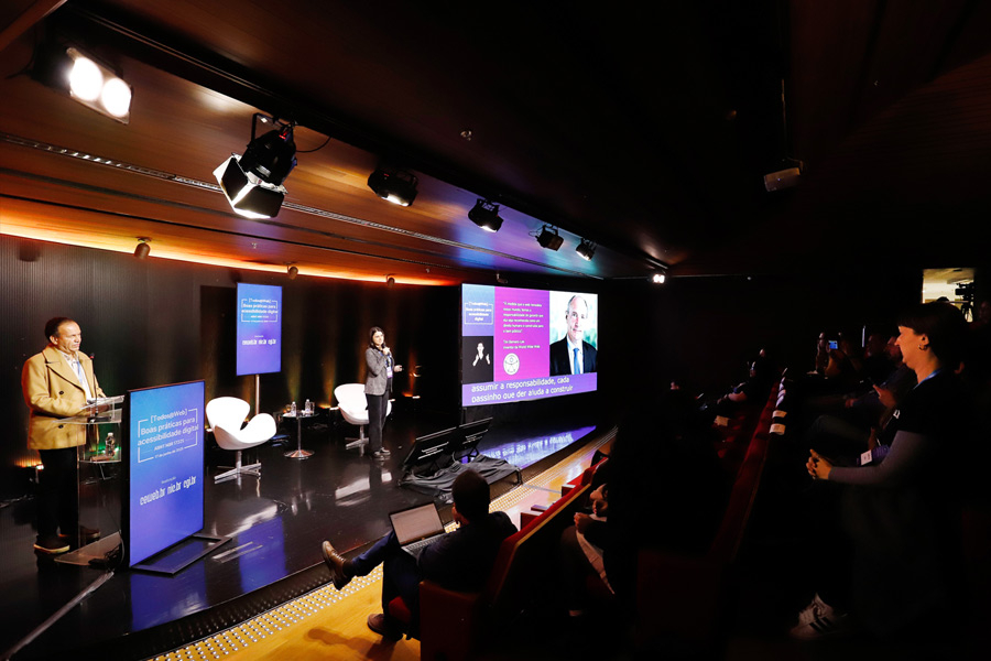 Foto de Cláudia Nascimento ministrando palestra no palco do auditório do Nic.br, à sua direita, está Sidney Tobias. À sua esquerda, tela com slides.
