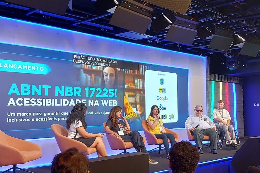 Cláudia Nascimento fala ao microfone em painel do Lançamento da norma ABNT NBR 17225. No painel, participam Amanda Marques, Simone Freire, Laércio Santana e um rapaz cadeirante.